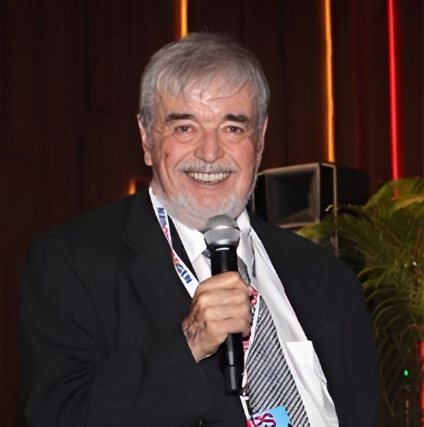 Smiling man in a suit holds a microphone, likely speaking at an event. Colorful background suggests a lively atmosphere.